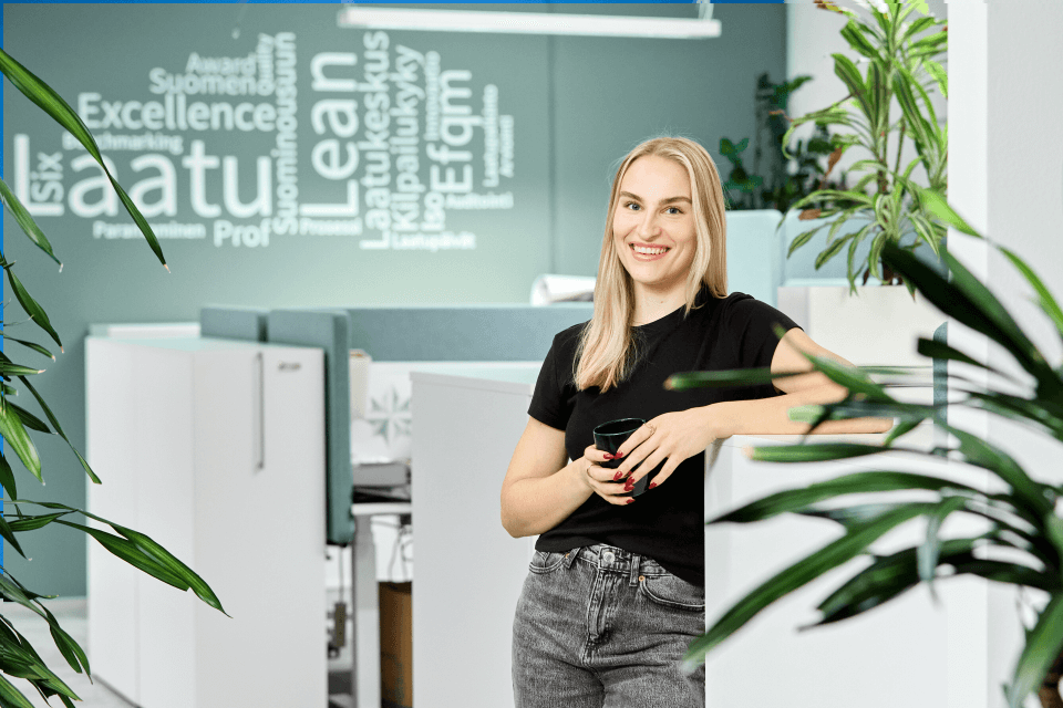 Hymyilevä nainen toimistoympäristössä. Etualalla viherkasveja, taustalla vaaleanvihreä seinä, jossa Excellence Finlandin osaamisalueita kuvaavia sanoja.